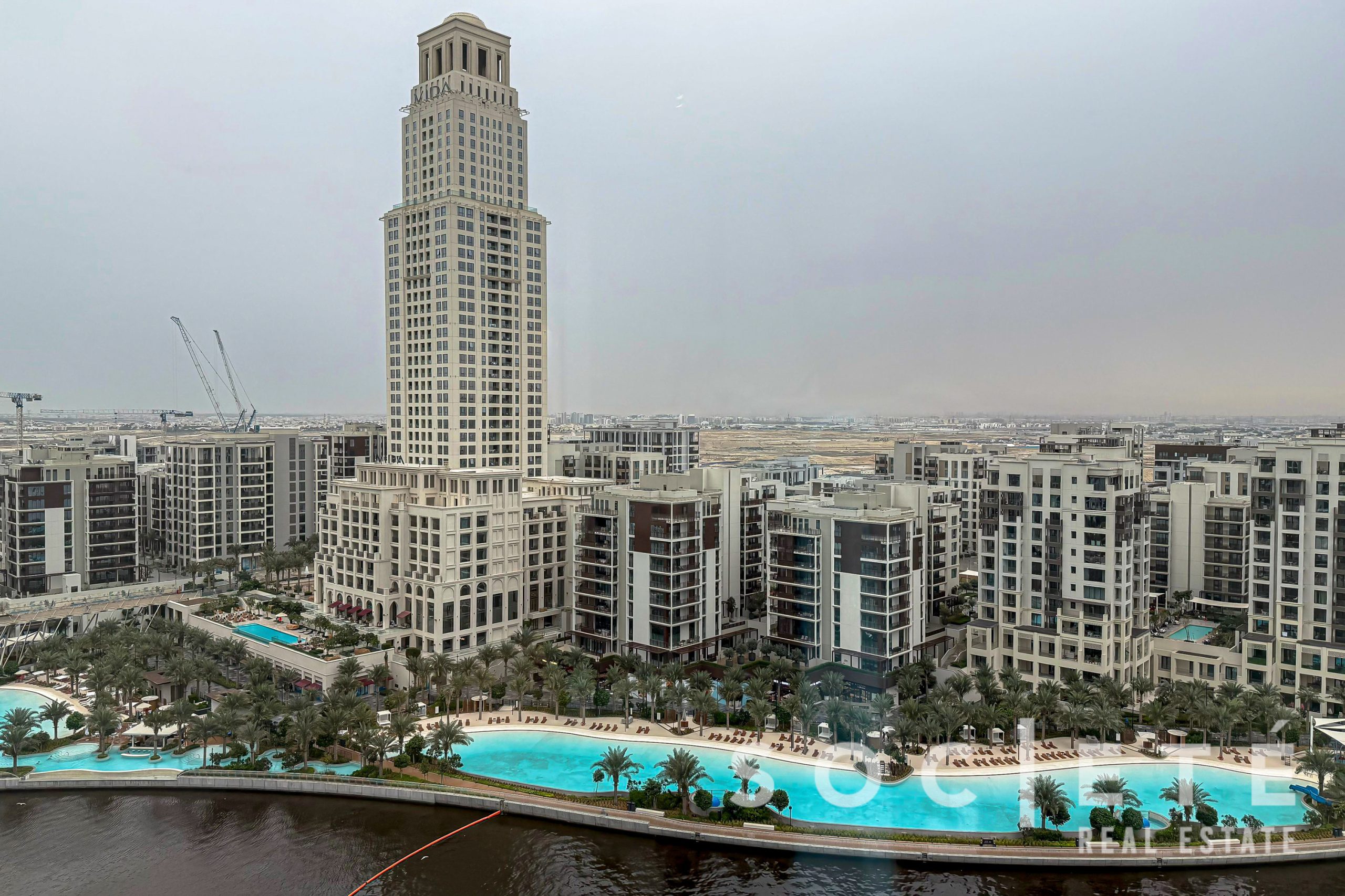 Tall beige skyscraper centered among white residential buildings, with a resort pool and palm trees in the foreground.