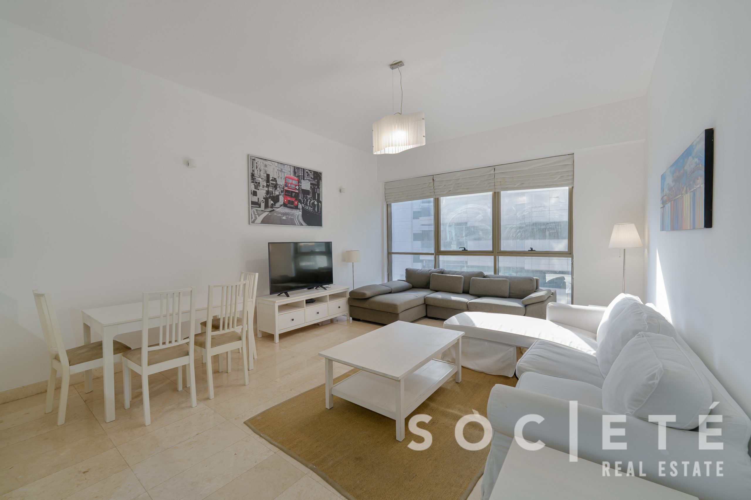 Bright living room with white dining set, TV stand, and a gray sectional sofa by large windows, beige rug under a white coffee table.