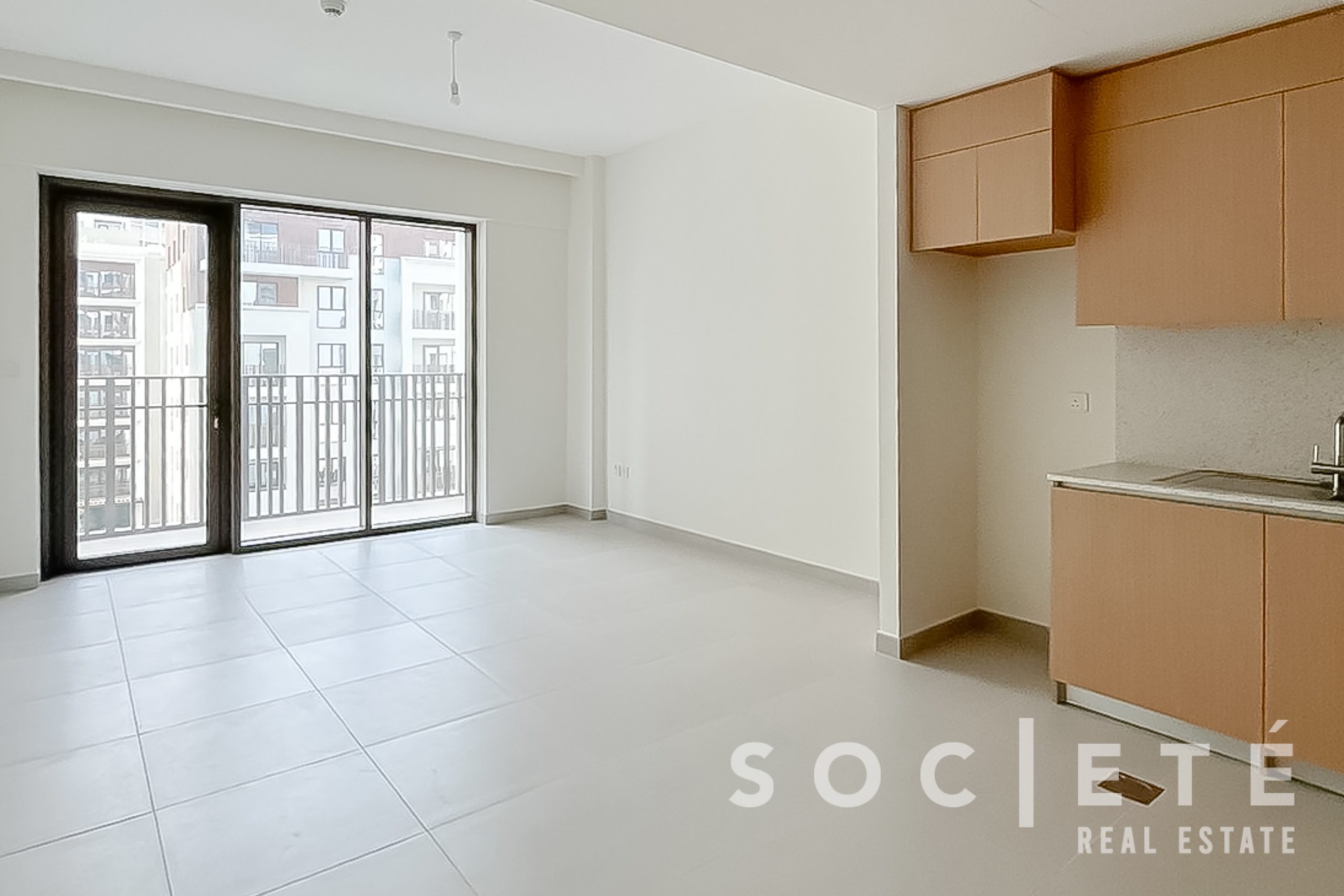 Open-plan living area with sliding glass doors to a balcony, white walls, and light gray tiled floor; brown kitchen cabinets on the right.