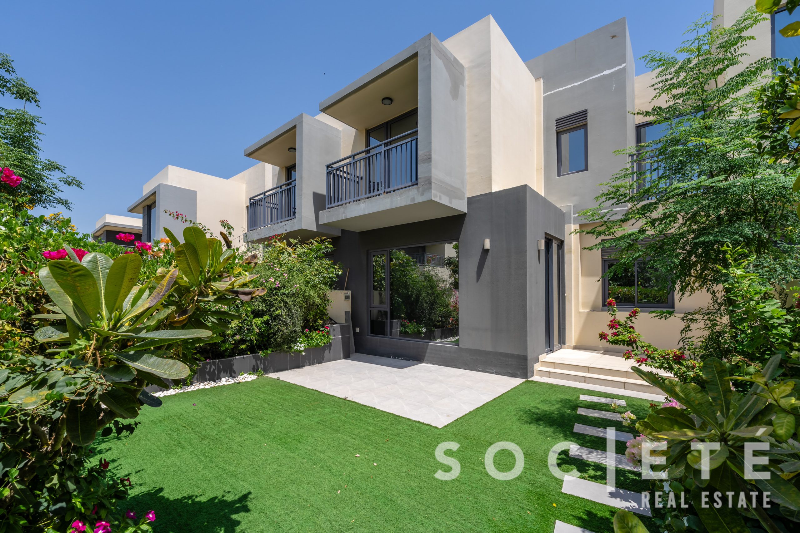 Modern apartment building with gray and beige walls, balconies, and lush tropical garden under a blue sky, view from ground level.