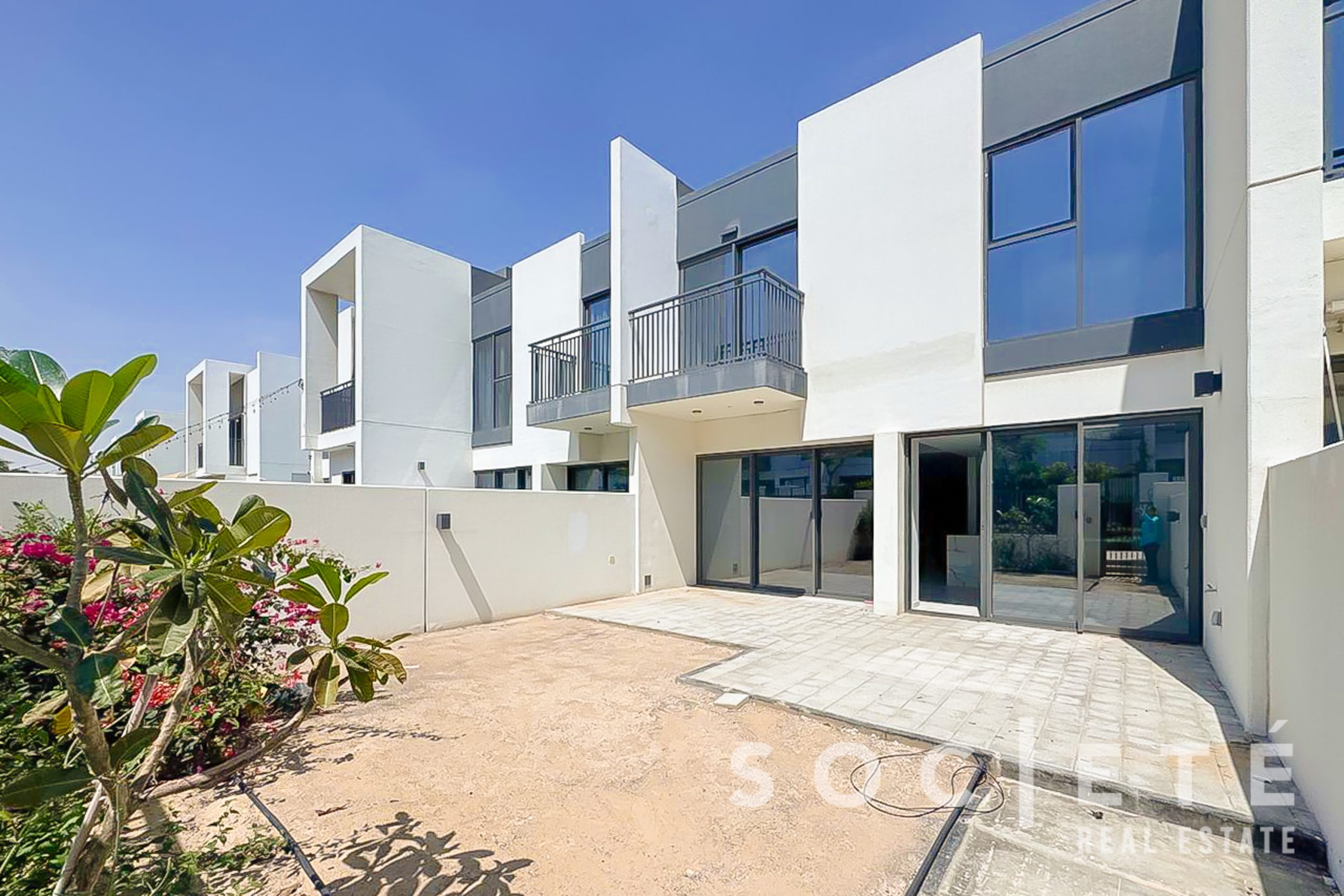 Modern white townhouse complex with large glass windows, balconies, and a paved entry courtyard under a clear blue sky.