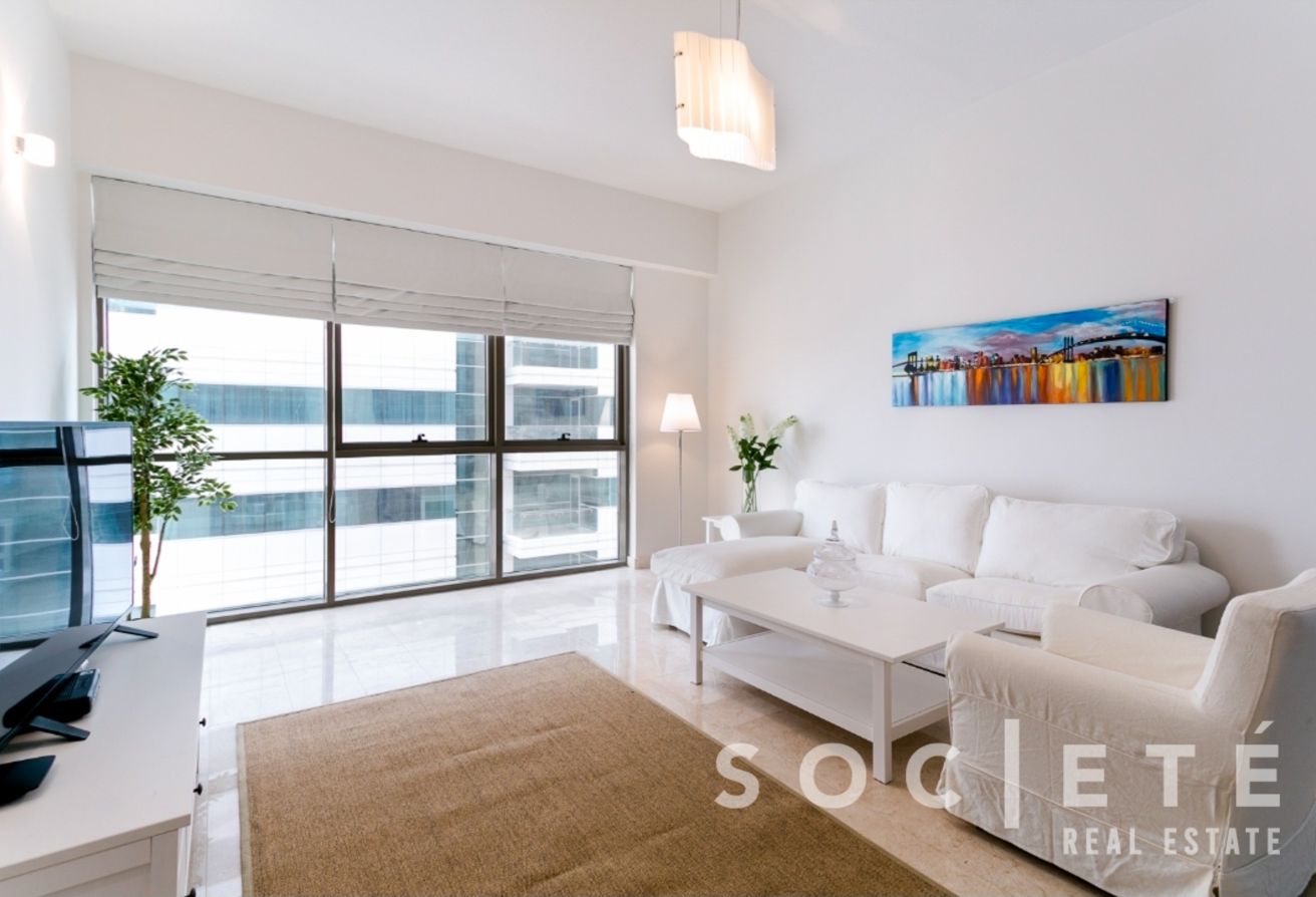 Bright modern living room with a white sofa, glass windows, and a cityscape view of tall buildings.