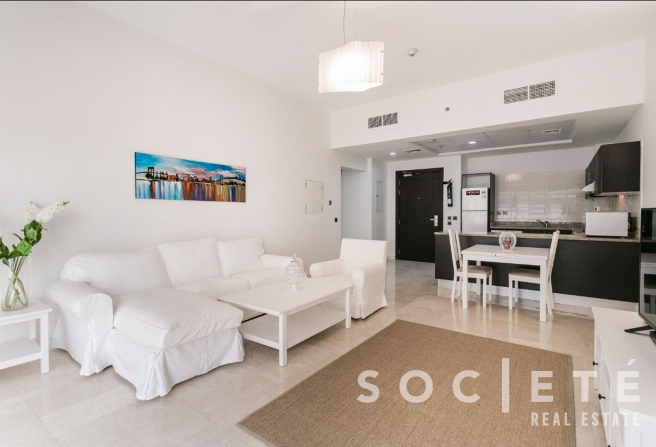 Bright, white living area with a white sofa and armchairs, coffee table, and a cityscape wall art; open kitchen in the background.