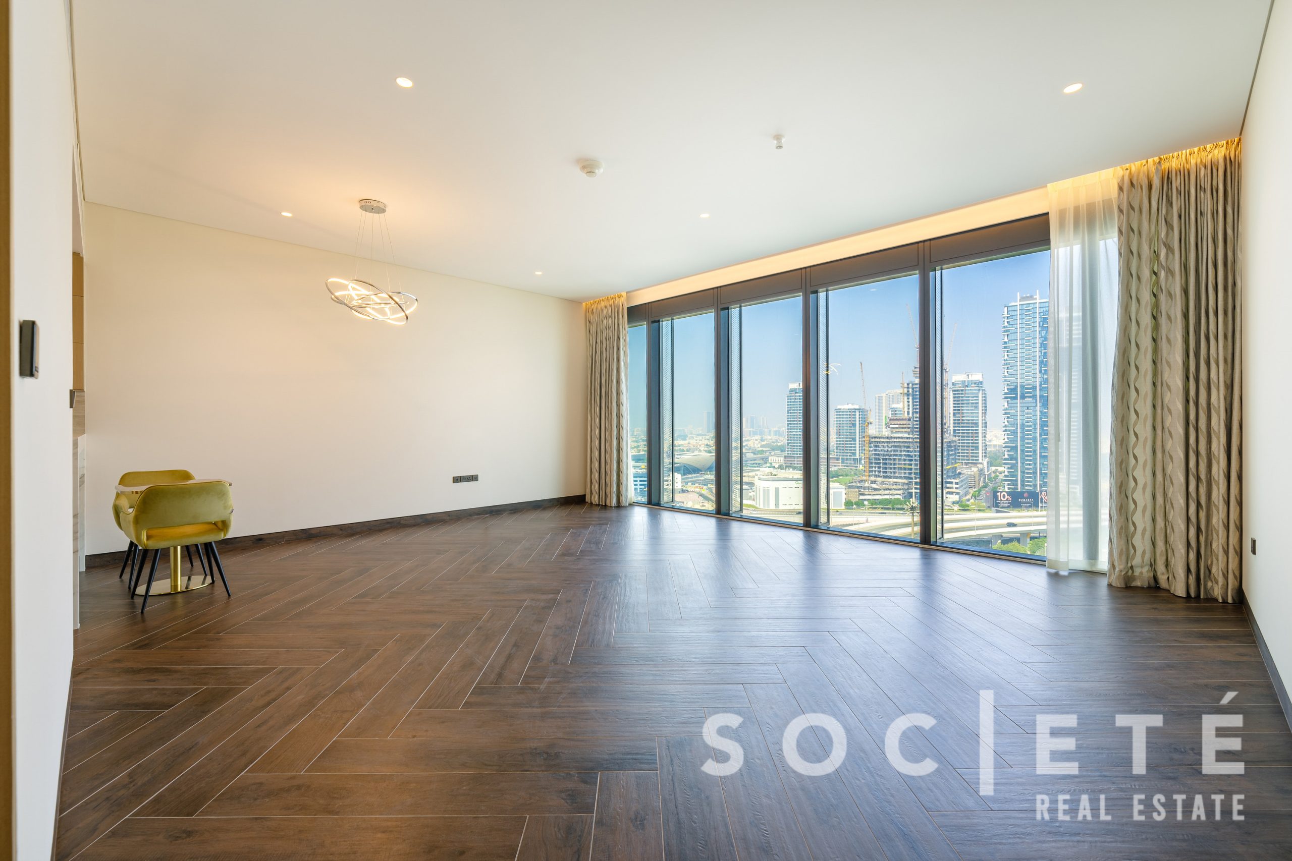 Spacious living room with dark wood floors, beige walls, and large floor-to-ceiling windows overlooking a city skyline; modern circular chandelier above a lime chair.