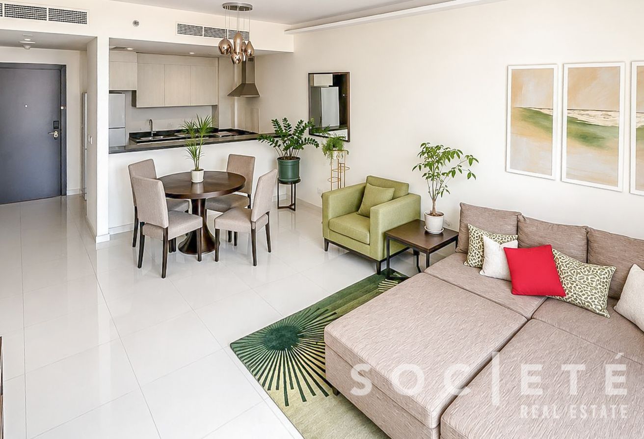 Open-concept living area with beige sectional sofa, round dining table, green armchair, and kitchen in the back; several potted plants.