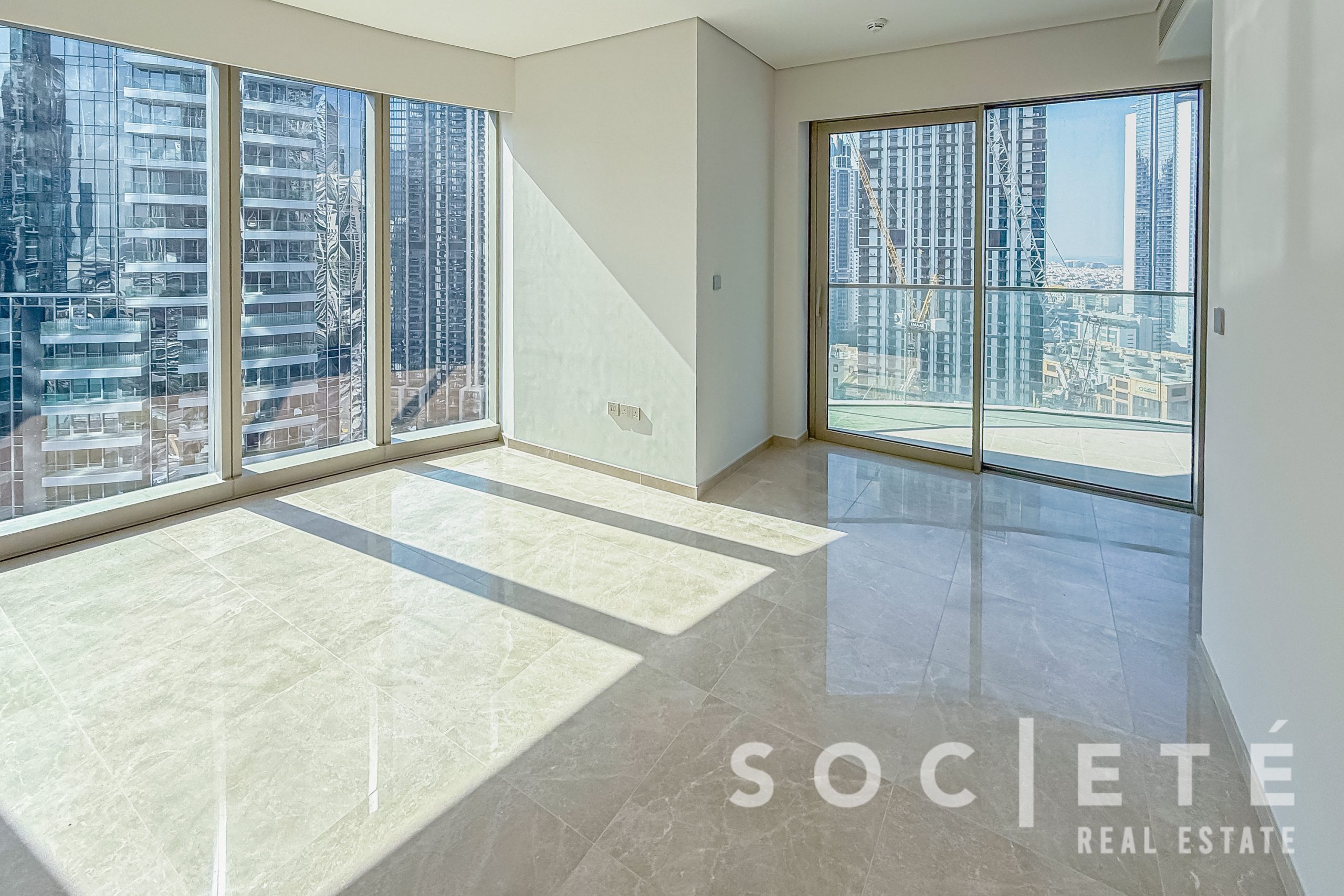 Sunlit empty living room with large floor-to-ceiling windows showing a city skyline of tall buildings and cranes outside.