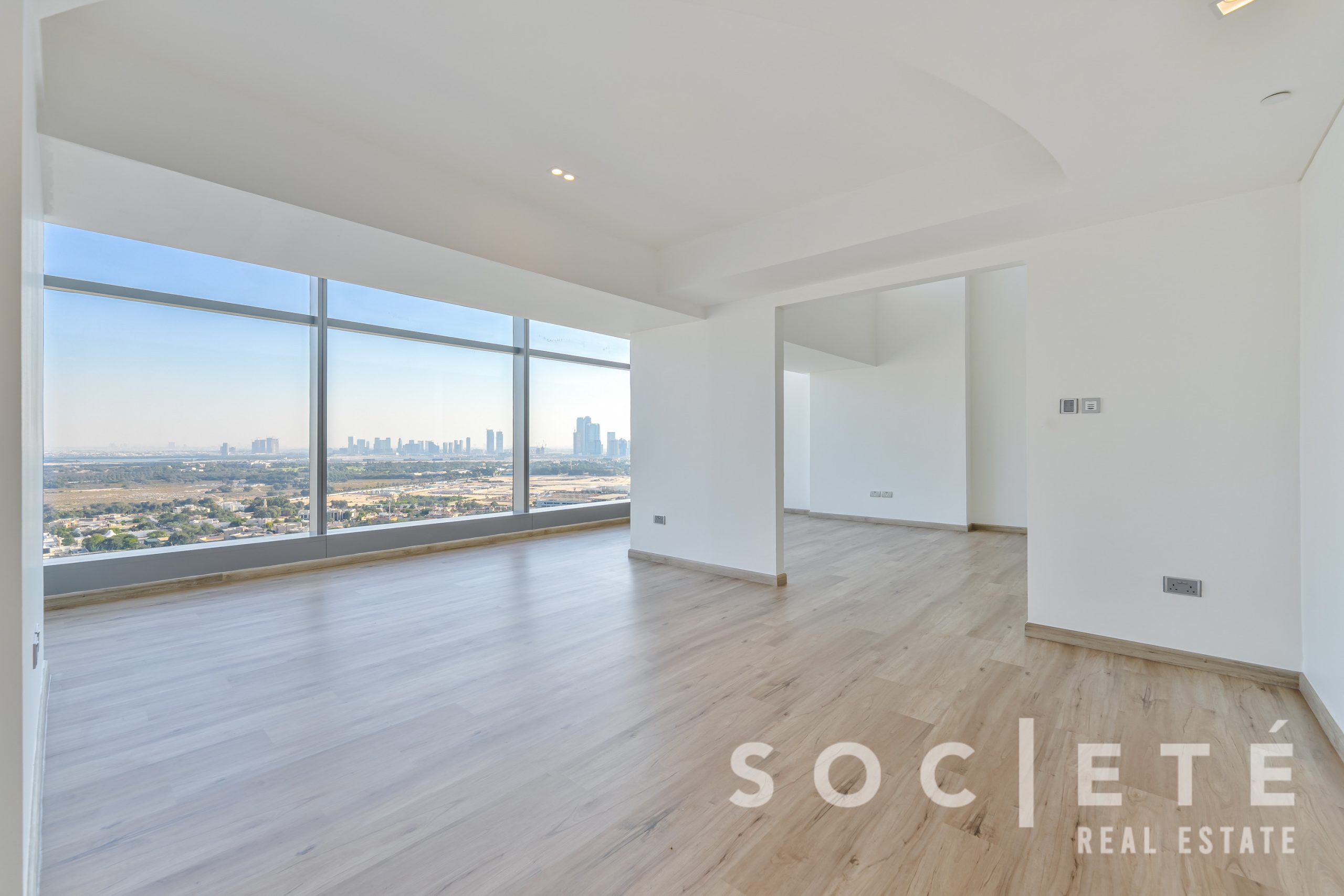 Bright empty living area with floor-to-ceiling windows and a city skyline view, light wood floors and white walls.
